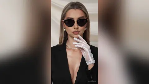 Elegant woman in a black blazer and white lace gloves wearing dark sunglasses in a chic studio setting.