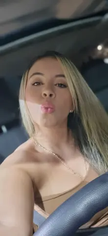 Blonde woman in a tan top making a kissy face while sitting in a car interior.