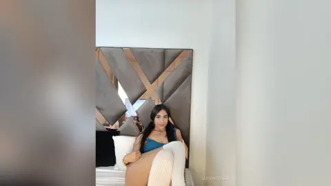 Young woman in a blue top and white knit sweater sitting on a bed in a modern bedroom.