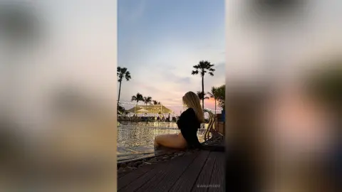 BiankaPerese sits on a wooden deck by a pool at sunset, wearing a black bodysuit or swimsuit with her blonde hair down.