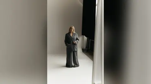 Blonde woman in a chic grey oversized suit standing in a minimalist bright studio setting.