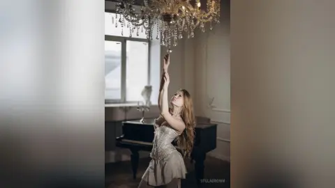 StelaBrown reaches up toward a crystal chandelier while wearing a light-colored corset and standing near a black piano.