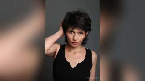 IveteDurand poses with one hand in her short dark hair, wearing a black tank top against a plain grey background.