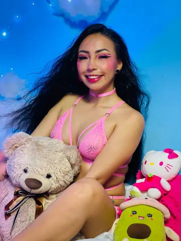 NathaliaPink smiles in a pink lace bikini and choker, sitting next to a large teddy bear and several plush toys against a blue background.