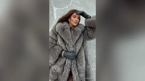 Elegant woman in a luxurious grey faux fur coat and black gloves posing against a textured wall.