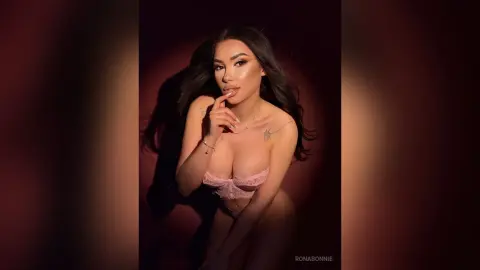 Glamorous woman in pink lace lingerie posing against a dark moody background with dramatic lighting.