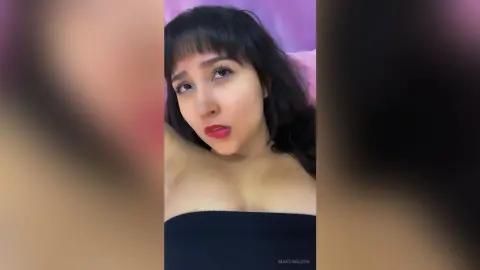 Young woman with dark bangs wearing a black tube top relaxing in a bedroom setting.