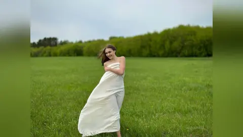 Smiling woman in a flowing white silk dress walking through a lush green meadow under a soft sky.