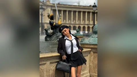 Stylish woman in a black beret and blazer posing by a classical fountain in a European city square.
