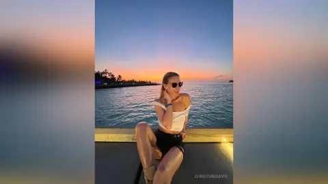 ChristineDavis poses by the ocean at sunset, wearing a white crop top and black shorts with her hair pulled back.