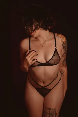 Isabella_Stewart poses in black polka-dot mesh lingerie, showing her tattoos and dark hair against a dark background.