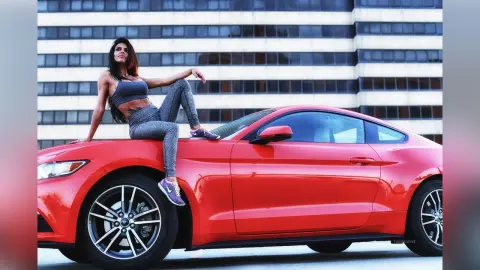 DrellaWolf — Athletic woman in grey gym wear sitting on a red Ford Mustang in front of a modern city building.