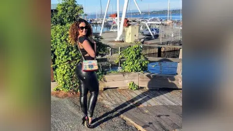 Smiling woman in black top and leather pants with a colorful crossbody bag at a sunny waterfront marina.