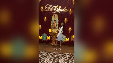 Young woman in a white top and denim skirt posing before a religious shrine with candles and a teal skull.