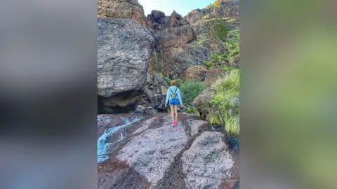 Adventurous female hiker in a blue shirt and shorts standing on rocky cliffs in a lush mountain canyon.