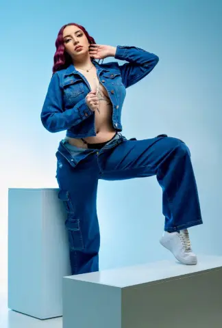Red-haired woman in a denim jacket and wide-leg jeans posing confidently against a blue studio background.