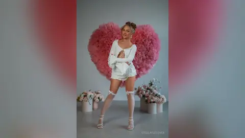 StellaFletcher stands in front of a large pink heart-shaped feather backdrop wearing a white lace teddy and white thigh-high stockings.