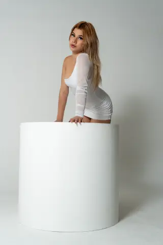 Blonde woman in a white long-sleeve mini dress posing elegantly against a minimalist white studio background.