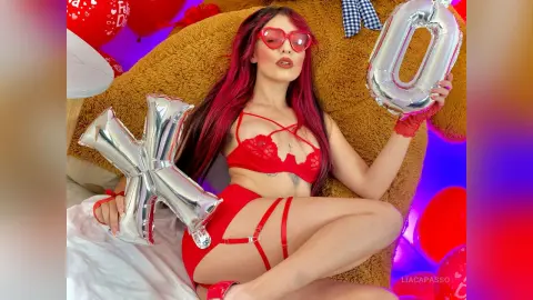 Woman with red hair in red lace lingerie relaxing with silver XOXO balloons in a cozy setting.