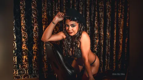 Glamorous woman in black lace lingerie posing against elegant gold patterned curtains in moody lighting.
