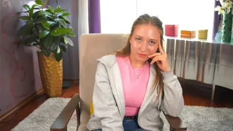 Young woman in a pink shirt and grey hoodie sitting relaxed in a cozy living room setting.