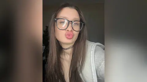Young woman with glasses and black choker making a playful pouty face in a casual indoor setting.