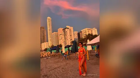Woman in an orange bikini standing on a beach at sunset with city skyscrapers in the background.