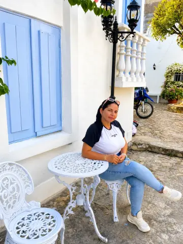 Victoria_Velvet sits at a white table outside wearing a white t-shirt and light blue jeans, with her hair pulled back and sunglasses on her head.