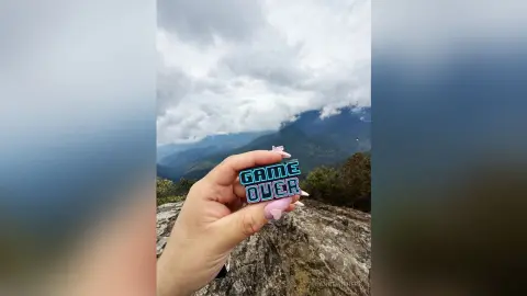 Hand holding a neon blue Game Over sign against a moody, cloudy mountain landscape.
