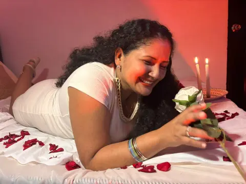 GenesisMontenegro smiles while lying on a bed covered in red rose petals, wearing a white dress and holding a white rose near lit candles.