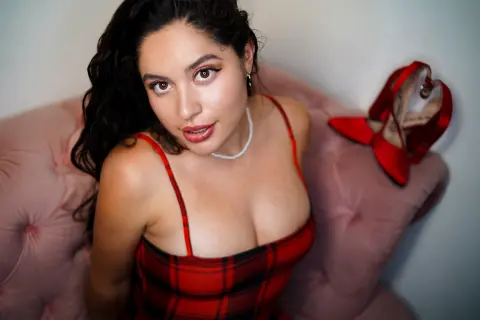 SharaThomas wears a red plaid dress and a pearl necklace while sitting on a pink velvet chair.