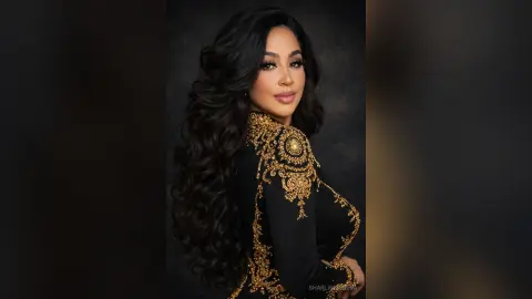 Elegant woman with long wavy hair wearing a black luxury gown with intricate gold embroidery in a moody studio setting.