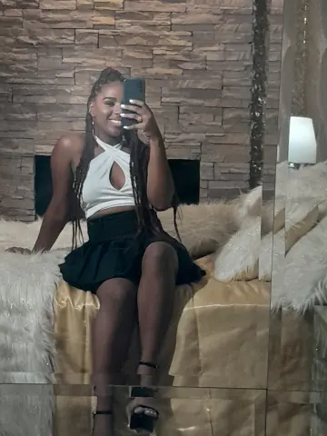 Smiling woman in a white halter top and black skirt sitting on a bed in a modern bedroom.