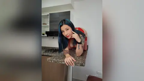 Blue-haired woman in a red top leaning on a kitchen counter in a modern home setting.