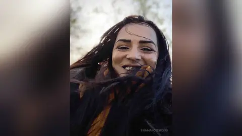 Smiling woman with long dark hair in a cozy mustard sweater outdoors in a natural setting.