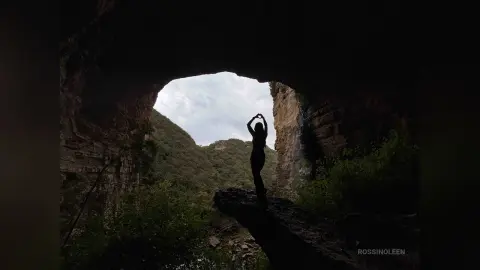 RossiNoleen stands in silhouette against a bright cave opening, raising her arms above her head.