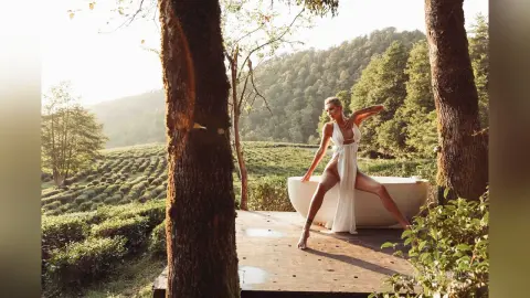 Elegant woman in a white flowing dress relaxing in an outdoor bathtub overlooking a lush green tea plantation.
