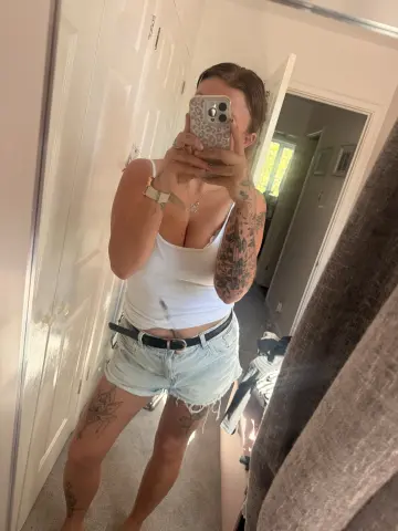 TattedBrat26 takes a mirror selfie wearing a white tank top and light denim shorts, showing off her many tattoos.