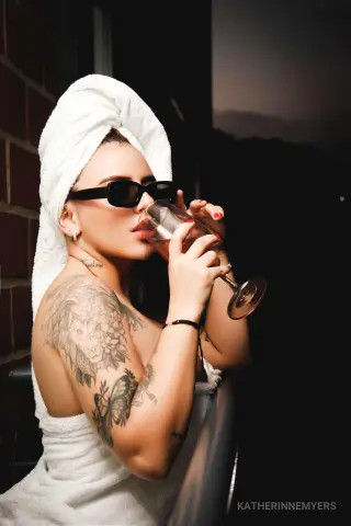 Tattooed woman in a white towel and sunglasses sipping wine on a balcony at night.