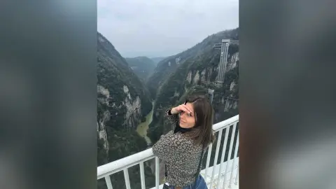 Young woman in a patterned sweater looking over a scenic mountain river valley from a high balcony.