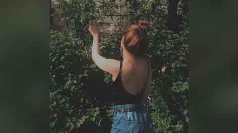 Young woman in a black tank top and denim jeans standing by lush green garden bushes in soft natural light.