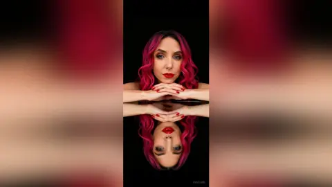Portrait of a woman with wavy pink hair and red lipstick posing against a dark background with a mirror reflection.