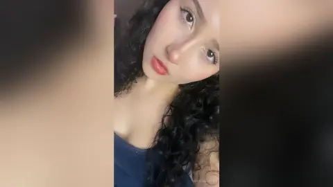 Young woman with curly dark hair and red lipstick posing for a close-up portrait in soft natural lighting.