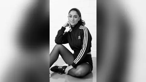 Smiling woman in black Adidas tracksuit and tights sitting against a white brick wall in a moody black and white portrait.