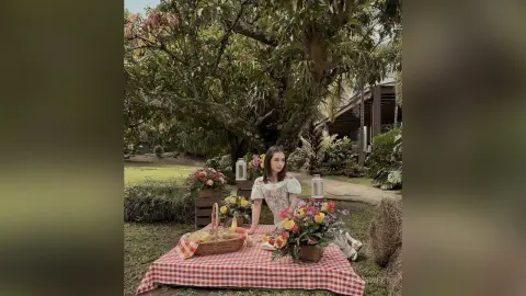LauriSweet sits behind a red and white checkered picnic blanket outdoors under a large tree, wearing a white lace top and looking at the camera.
