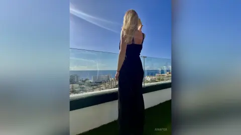 AmaFox — Blonde woman in a black evening dress overlooking a sunny coastal city view from a luxury balcony.