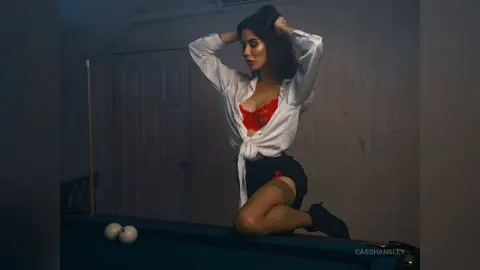 Moody portrait of a woman in a white shirt and red lace bra sitting on a pool table in a dimly lit room.