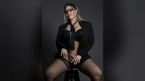 Confident woman in a black blazer, tie, and lace lingerie sitting against a dark studio background.