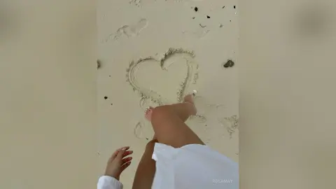 RosaMay lies on a sandy beach, showing her bare legs and feet near a heart drawn in the sand.