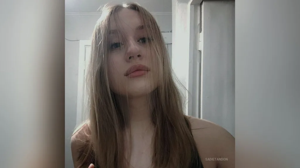 Close-up portrait of a blonde woman with natural makeup looking calmly at the camera in soft indoor lighting.